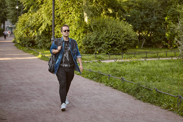 Young man is walking in public park. Caucasian tourist in sunglasses and with rucksack walks outdoors. - Φωτογραφία, εικόνα