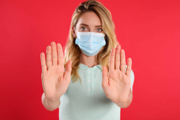 Woman in protective face mask showing stop gesture on red background, focus on hands. Prevent spreading of coronavirus - 写真・画像