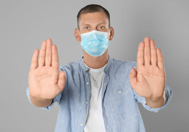 Man in protective mask showing stop gesture on grey background. Prevent spreading of coronavirus - 写真・画像