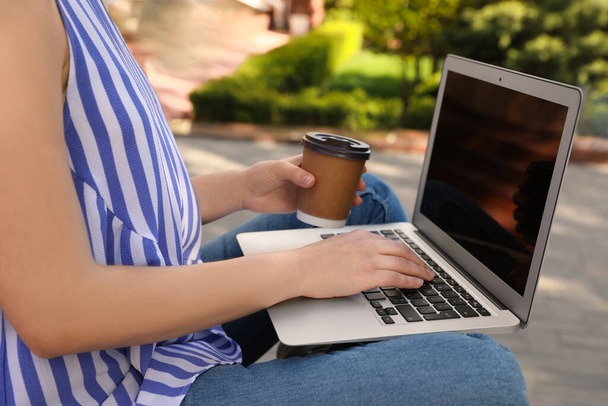 Young woman with paper cup of coffee working on laptop outdoors, closeup - 写真・画像