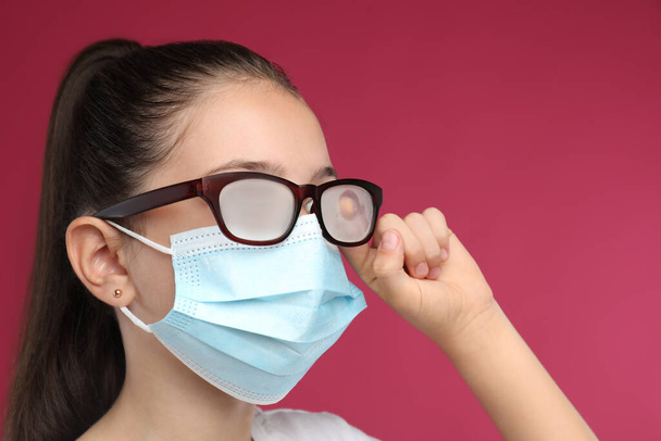 Little girl wiping foggy glasses caused by wearing medical face mask on pink background. Protective measure during coronavirus pandemic - Фото, изображение