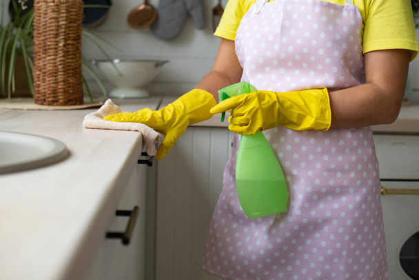Уборка, уборка дома. Гигиена и чистота на кухне. Девушка в жёлтых резиновых перчатках и фартуке снимает. Рука вытирает поверхность стола и держит спрей - Фото, изображение