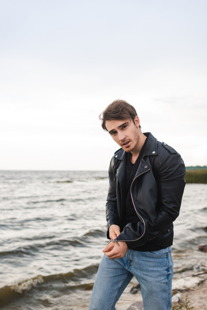 Hombre joven en jeans y chaqueta de cuero mirando a la cámara en la playa  - Foto, Imagen