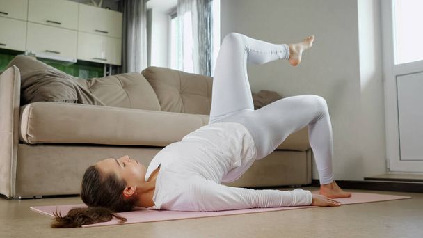 woman does sports exercises lying on pink mat near couch - Foto, afbeelding