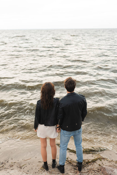 Back view of man in leather jacket standing near girlfriend on beach near sea  - Fotó, kép