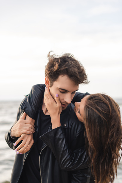 Young woman touching cheek and embracing boyfriend in leather jacket near sea - Photo, Image
