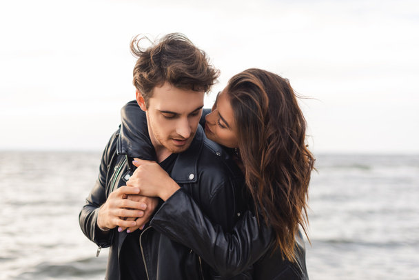 Young brunette woman hugging boyfriend in black leather jacket near sea - Foto, Imagem