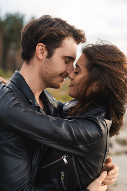 Young man kissing brunette girlfriend in leather jacket outdoor - Photo, Image
