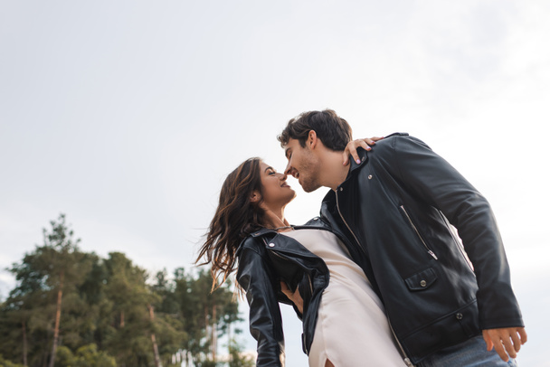 Baixo ângulo vista do homem na jaqueta de couro beijando namorada no vestido ao ar livre  - Foto, Imagem
