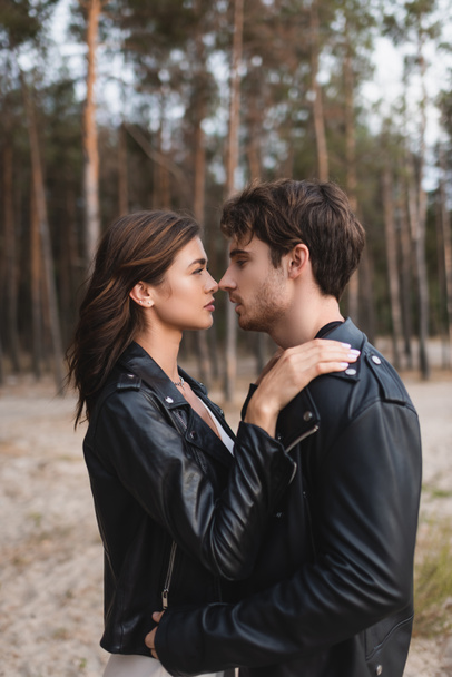 Side view of brunette woman in leather jacket embracing boyfriend in forest  - Photo, Image