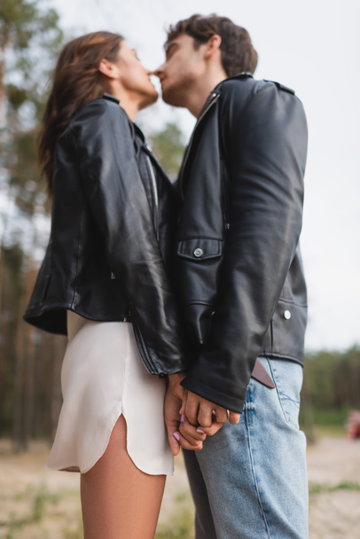 Low angle view of young couple in leather jackets holding hands and kissing in forest  - Photo, Image