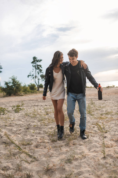 Couple in leather jackets embracing while walking with bottle of wine on sandy beach  - Foto, imagen