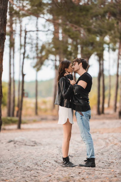 Side view of man in leather jacket kissing brunette girlfriend on sand in forest  - Foto, afbeelding
