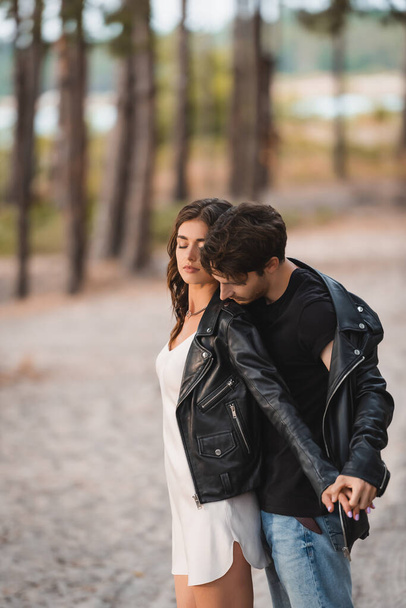 Man holding hand of brunette girlfriend in leather jacket in forest - Фото, изображение