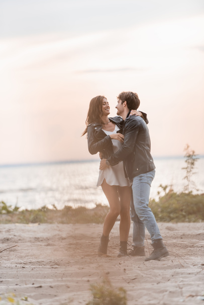 Selective focus of young couple in leather jackets hugging on beach at dawn  - Photo, Image