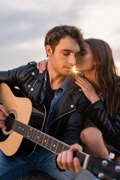 Selective focus of woman kissing boyfriend with acoustic guitar at sunset  - Fotografie, Obrázek
