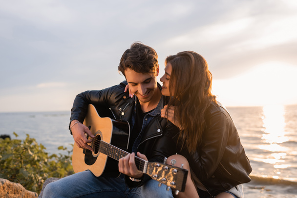 Concentration sélective de la femme en veste en cuir embrassant petit ami jouant de la guitare acoustique pendant le coucher du soleil sur la plage  - Photo, image