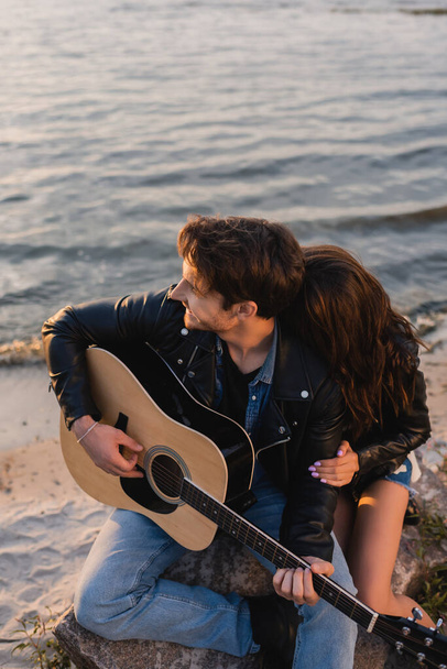 Frau umarmt Freund, der am Strand Akustikgitarre spielt  - Foto, Bild