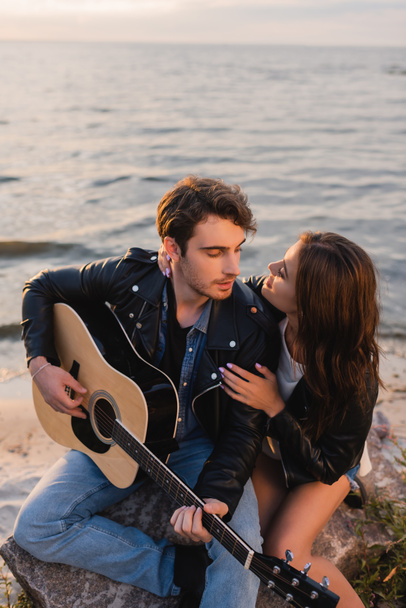 Giovane donna che abbraccia fidanzato con chitarra acustica in spiaggia la sera  - Foto, immagini