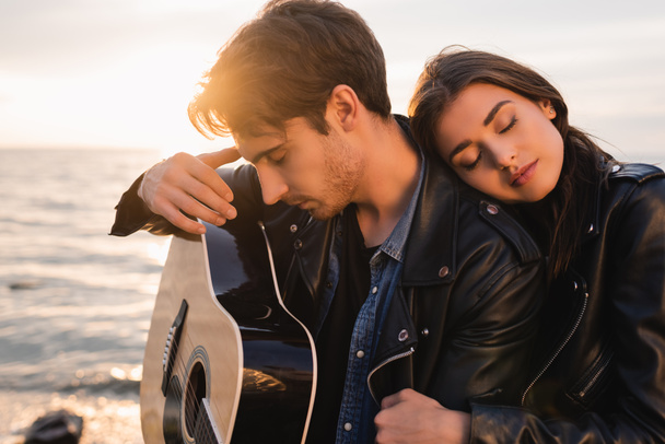 Młody mężczyzna trzyma gitarę akustyczną w pobliżu kobiety na plaży o zachodzie słońca  - Zdjęcie, obraz