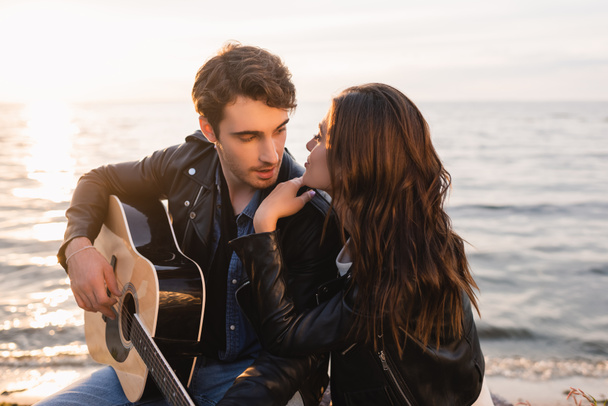 Young woman looking at boyfriend with acoustic guitar on beach at sunset - Photo, Image
