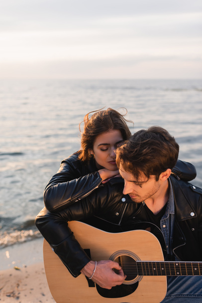 Young woman hugging boyfriend with acoustic guitar on beach at sunset  - Photo, Image