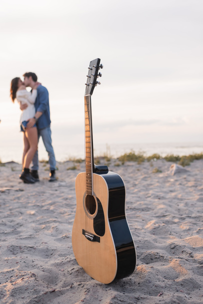 Selective focus of acoustic guitar on sand and young couple kissing on beach at sunset  - Foto, Imagem