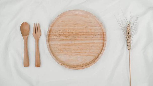Πιάτο ξύλινο, κουτάλι ξύλινο και πιρούνι ξύλινο με Barley ξηρό λουλούδι σε λευκό πανί. Απομίμηση μαγειρικών σκευών. Κάτοψη της ρύθμισης του πίνακα σε λευκό φόντο - Φωτογραφία, εικόνα
