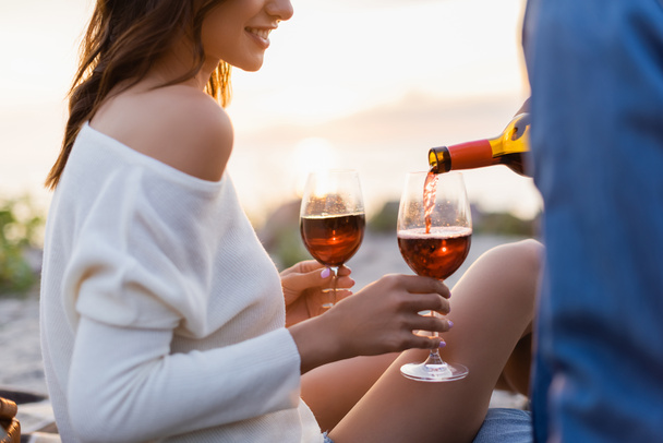 Cropped view of woman holding glasses while boyfriend pouring wine on beach at evening - Foto, Imagen