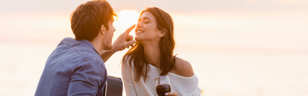Panoramic shot of man with acoustic guitar touching nose of girlfriend with glass of wine near sea - Photo, Image