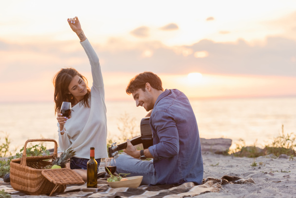 ビーチでのピクニック中にアコースティックギターを演奏するボーイフレンドの近くにワインのガラスを保持している女性の選択的な焦点  - 写真・画像