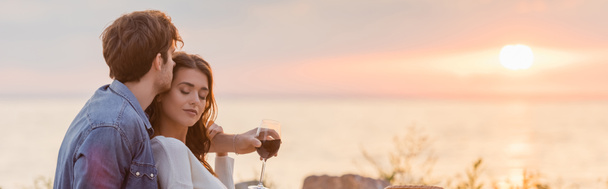 Panoramaaufnahme einer Frau mit einem Glas Wein, die ihre Freundin am Strand bei Sonnenuntergang umarmt  - Foto, Bild