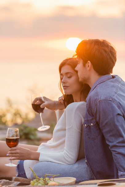 Selective focus of man embracing girlfriend with glass of wine near acoustic guitar on beach  - Fotoğraf, Görsel