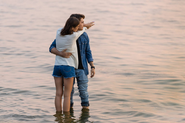 Young couple embracing while standing in water of sea at evening  - Foto, imagen