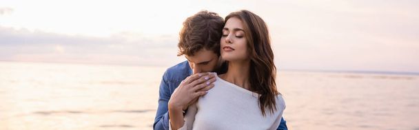 Panoramic shot of man touching brunette girlfriend with closed eyes near sea at sunset  - Photo, Image