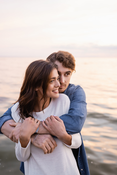 Man embracing girlfriend and looking away near sea at sunset  - Photo, Image