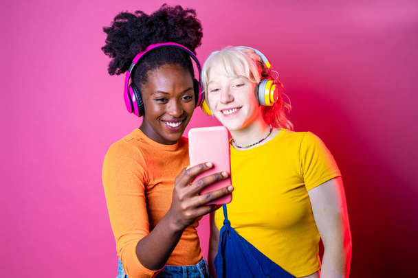 Dwóch młodych, wielonarodowych przyjaciół noszących na różowym tle selfie - Izolowane, zróżnicowane kobiety streamujące na żywo za pomocą smartfona - vlogging, streaming, social networks concept - Zdjęcie, obraz
