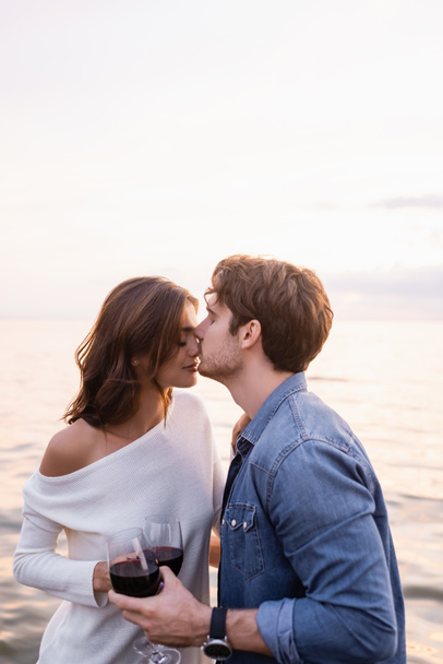 Selective focus of young man kissing nose of girlfriend with glass of wine near sea  - Photo, Image
