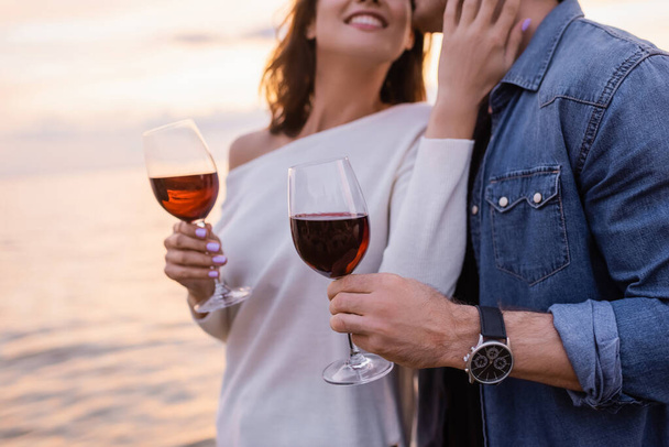 日没時に海の近くでワインのガラスを保持しながら彼氏に触れる女性の作物のビュー  - 写真・画像