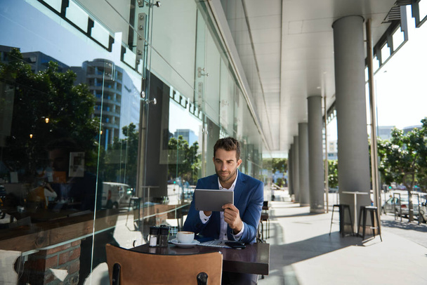 若いビジネスマンがコーヒーを飲みながら、カフェテーブルに座っている間にデジタルタブレットを使用しています。 - 写真・画像