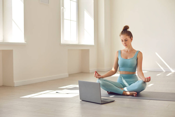 Young yoga coach sitting cross legged in front of laptop giving online lesson or hosting webinar - Foto, afbeelding