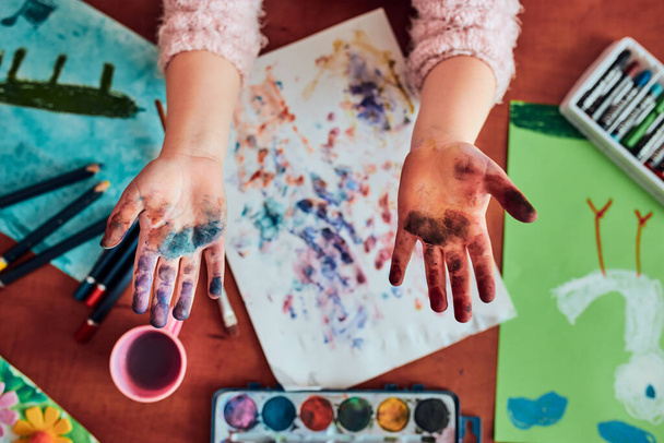 Pieni tyttö esikoululainen näyttää maalatut värikkäät kädet. Lapsella on hauskaa tehdä postimerkkejä paperiarkille maalatuilla käsillä luokkahuoneen taidekurssin aikana. - Valokuva, kuva