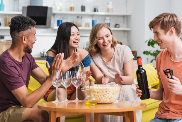 young man holding bottle of red wine near excited multicultural friends - Photo, Image