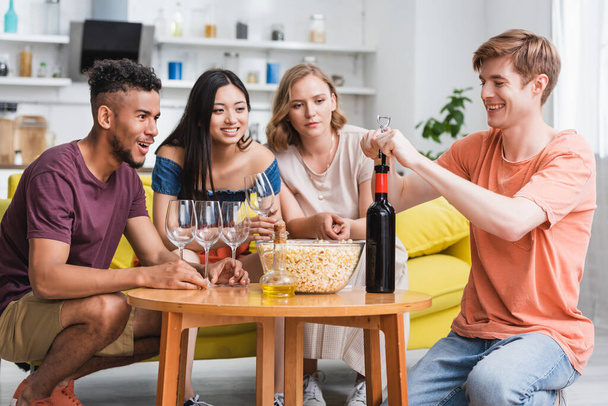 young man opening bottle of red wine near joyful multicultural friends - Foto, afbeelding