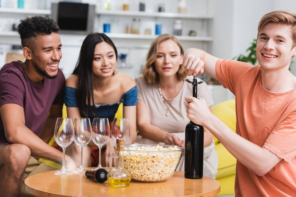 young man opening bottle of red wine near multicultural friends - Photo, Image