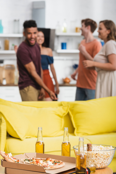 selektivní zaměření piva a pizzy na stůl v blízkosti žluté pohovky a multietnické přátele tanec na pozadí - Fotografie, Obrázek