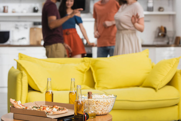 selective focus of pizza and beer on table near yellow sofa and multicultural friends dancing on background - Фото, изображение