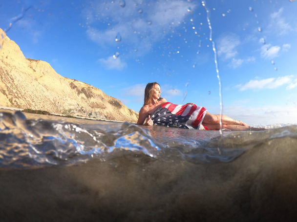 Una hermosa modelo morena posa desnuda con una bandera americana en la playa  - Foto, imagen