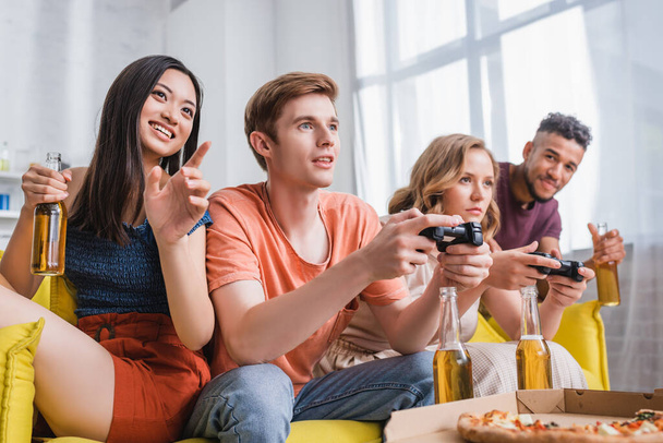 KYIV, UKRAINE - JULY 28, 2020: excited asian woman holding beer and pointing with finger near friends playing video game - Zdjęcie, obraz