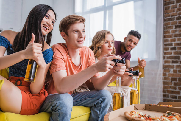 KYIV, UKRAINE - JULY 28, 2020: excited asian woman holding beer and showing thumb up near friends playing video game - Photo, Image